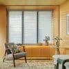 Layered horizontal sheer-and-opaque roller shades covering three tall window panels, partially closed and filtering soft daylight; set in a warm plywood-walled sitting nook with armchair, side table, lamp, and plant.