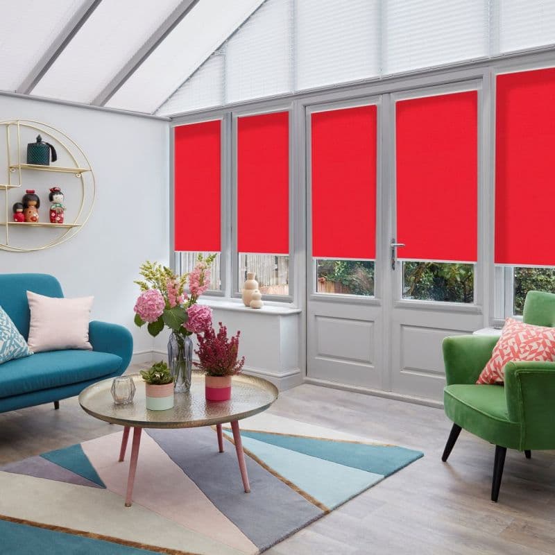 Four red roller blinds lowered over tall glazed windows and door to just above sills, filtering daylight and obscuring the view in a bright conservatory with teal sofa, green armchair.