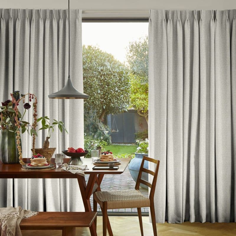 Floor-length grey pinch-pleat curtains, textured and hung on a concealed track, parted at center, admitting bright morning light, framing a sunlit backyard beyond a breakfast table with wooden chairs.