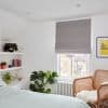 Grey fabric roman blind lowered over a sash window, filtering daylight; white bedroom with plants, wicker chair, radiator and a framed yellow print.
Transcription: THE SUN SHINES HERE EVERY DAY. MOSTLY.