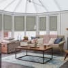 Pleated honeycomb blinds pulled down across tall conservatory windows, filtering soft daylight; the sunroom houses a beige sofa with chunky pink throw, wooden coffee table and a blue patterned rug.