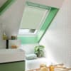Pale cream pleated skylight blind, mounted in a sloped window and mostly lowered with a small gap, filtering daylight; bright bathroom with green trim, white herringbone tiles, sink and tub.