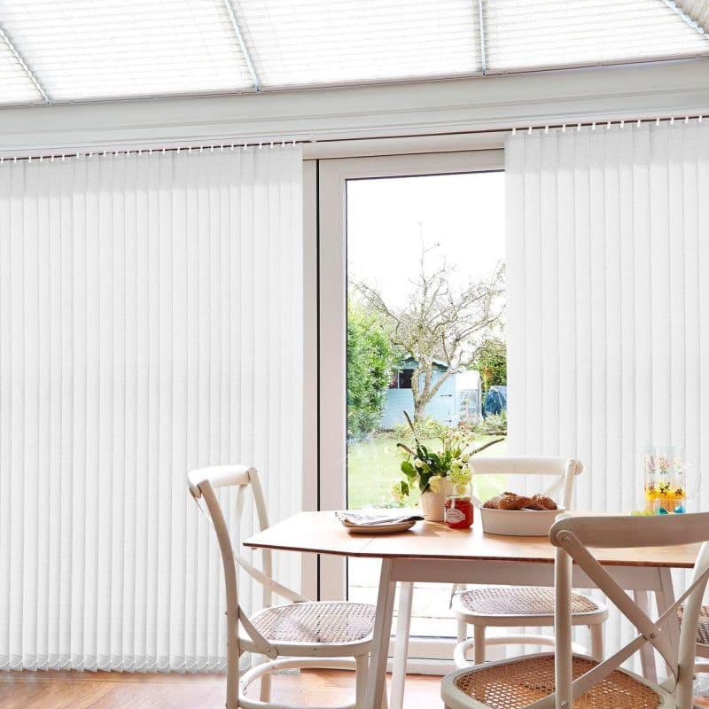 White vertical fabric blinds covering a floor-to-ceiling sliding glass door, mostly closed but parted centrally to reveal a sunlit garden; dining table and chairs in a bright breakfast nook.