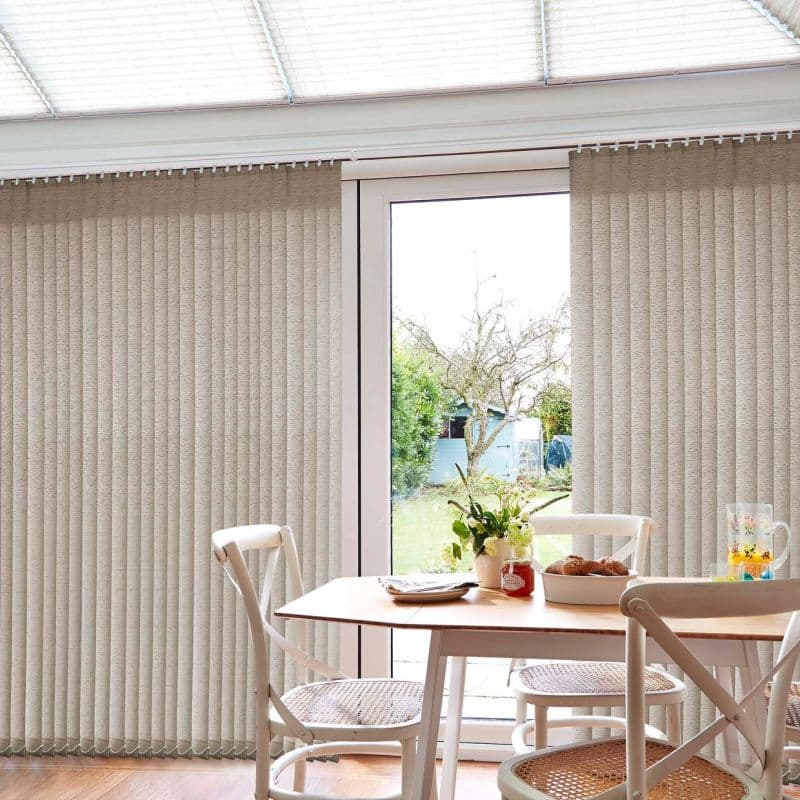 Beige textured vertical blinds, ceiling-mounted and floor-length, panels parted at center to reveal a glass patio door; filtering soft daylight into a small dining area with wooden table, chairs, vase.