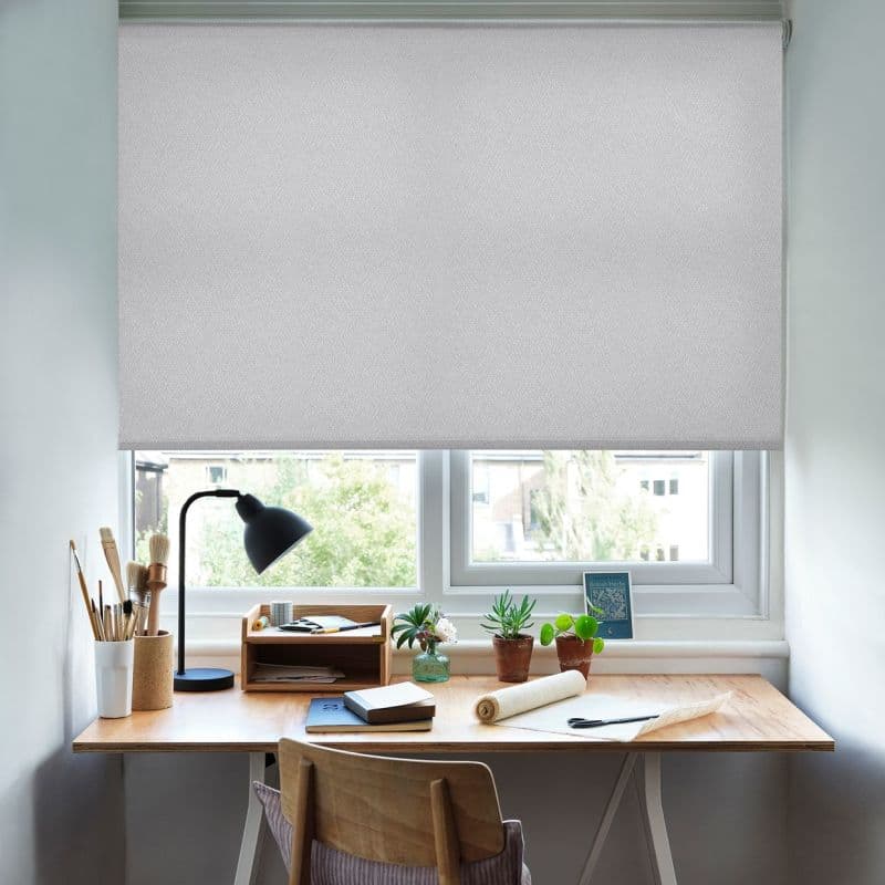 Light-grey textured roller blind lowered halfway, filtering daylight over a double window; a wooden desk beneath holds a lamp, brushes, small potted plants and notebooks in a bright home workspace.