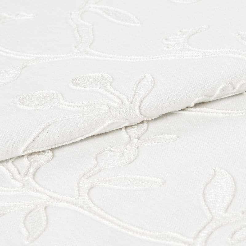 White embroidered fabric folded and overlapping, showing raised floral vine stitching and textured weave; close-up studio view with soft, even lighting emphasizing subtle shadows and fabric detail.