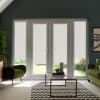 French glass doors covered by lowered white roller blinds, letting a strip of garden light through, centered in a living room with green armchairs, glass coffee table and chevron rug.
