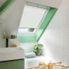 White pleated skylight blind, partially lowered and filtering soft daylight, in a sloped bathroom with mint-green window trim, white herringbone tiles, sink and bathtub with plant and bath tray.