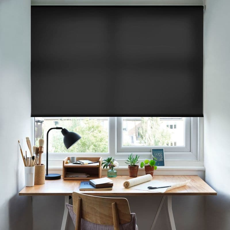 Black roller blind, fully lowered and opaque, blocking most light; installed over a double-pane window. Below, a wooden desk workspace with lamp, art brushes, notebooks and small potted plants.
