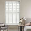 White plantation shutters—four hinged panels with horizontal louvers—cover a tall window; closed, filtering daylight into a neutral bedroom with upholstered bed, bedside table, lamp, armchair, and flowers.