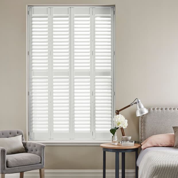White plantation shutters—four hinged panels with horizontal louvers—cover a tall window; closed, filtering daylight into a neutral bedroom with upholstered bed, bedside table, lamp, armchair, and flowers.