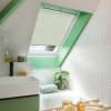 A cream pleated skylight blind, mostly lowered and filtering daylight through a slanted roof window; attic bathroom with bright green reveal, white herringbone tiles, sink, tub, plant and bath tray.