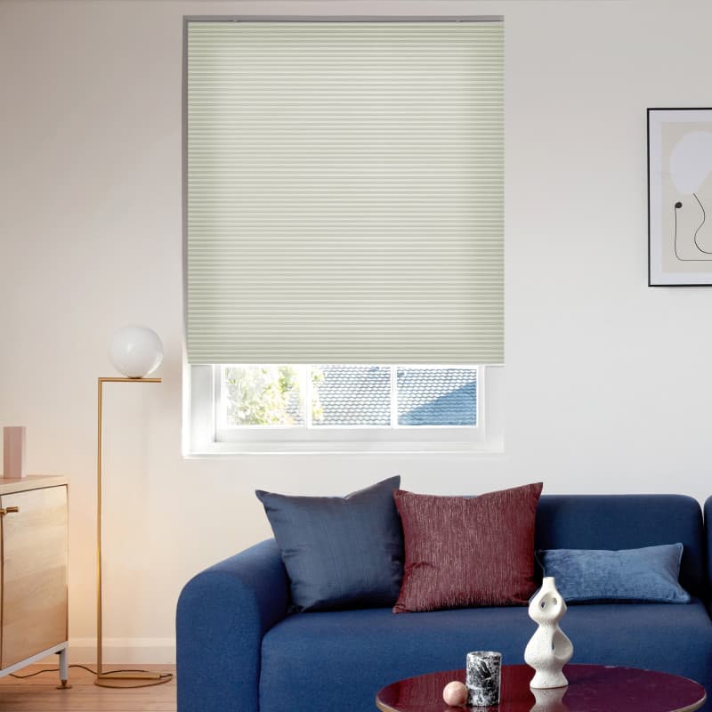 Light-green pleated cellular blind, fully lowered with horizontal ribs, filtering daylight through a small bottom gap — covering a living-room window above a blue sofa with pillows, lamp, and coffee table.