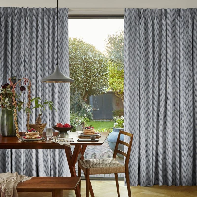 Gray patterned, floor-to-ceiling curtains, parted centrally and filtering soft daylight; a wooden dining table set with pancakes and fruit, pendant lamp above, chair and sunlit garden visible outside.