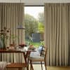 Beige pinch-pleat floor-length curtains framing a central opening, partially open and filtering daylight; in a dining room beside wooden table set for breakfast, pendant light and sunlit garden beyond.