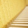 Vertical fabric blinds arranged along a side wall, scalloped bottoms linked by a bead chain, filtering strong sunlight into parallel stripes across a floor beside yellow leaf‑pattern wallpaper.