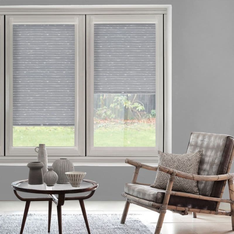 Grey roller blinds with subtle horizontal white lines on dual casement windows—left fully lowered, right mid-height—filtering daylight. Softly lit living room with rattan armchair, side table, and backyard lawn visible.