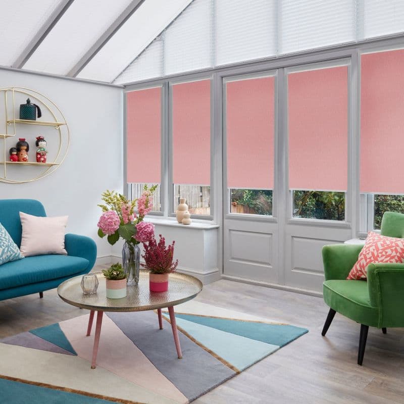 Pink fabric roller blinds, lowered over tall conservatory windows and doors with triangular skylights, partially closed and filtering daylight; cozy sitting room with colorful sofas, coffee table, and flowers.
