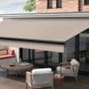 Retractable fabric awning, beige and extended from the fascia with a black roller, shading a glass sliding-door wall; outdoor patio with wicker chairs, round coffee table, built-in sofa, soft daylight.