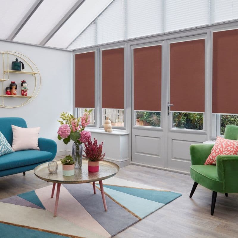 Brown roller blinds, lowered across tall French doors and windows, covering upper panes while leaving narrow lower glimpses; blocking daylight and providing privacy in a bright conservatory.