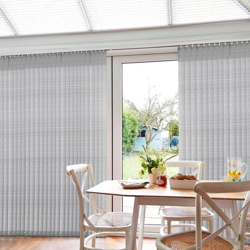Light-gray vertical blinds cover a sliding patio door, parted centrally to reveal the glass and filter daylight; bright dining area with wooden table, white chairs, flowers and bread, garden view.