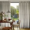 Pleated beige floor-to-ceiling curtains, parted centrally to frame a glass door, filtering soft sunlight; dining table set with pancakes and fruit, pendant lamp overhead, view of a lush backyard.