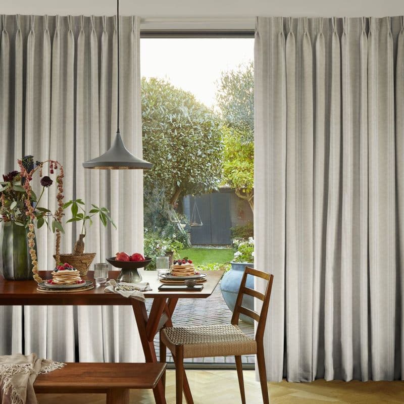 Pleated beige floor-to-ceiling curtains, parted centrally to frame a glass door, filtering soft sunlight; dining table set with pancakes and fruit, pendant lamp overhead, view of a lush backyard.