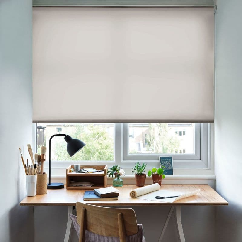 Beige fabric roller blind lowered halfway, blocking upper view and softly diffusing daylight. Below, a wooden desk workspace with lamp, art brushes, notebooks and small potted plants beside the window.