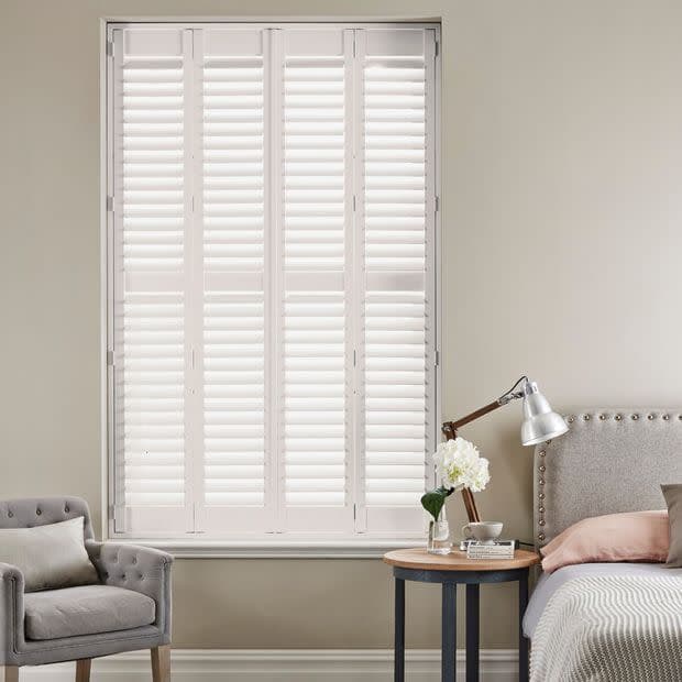 White plantation shutters covering a tall central window, louvered panels closed and filtering soft daylight; bedroom setting with upholstered bed, bedside table and lamp, plus an armchair against neutral walls.