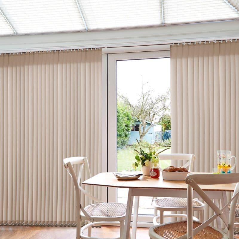 Beige vertical blinds cover a sliding glass door, parted at center to reveal a garden and admit soft daylight into a bright dining nook with wooden table and chairs.