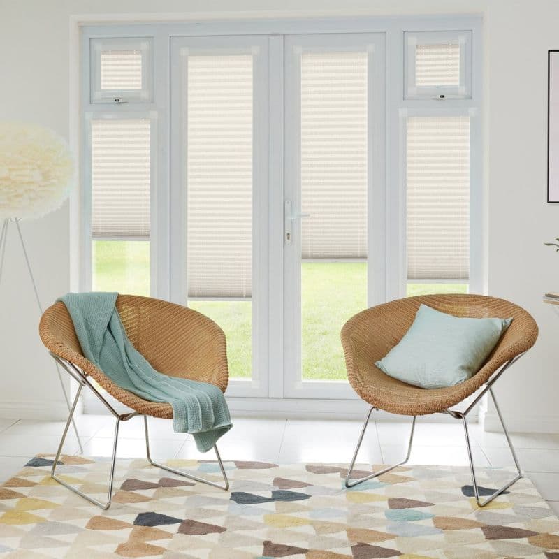 Light beige pleated blinds cover French doors and sidelights, lowered to mid-height, filtering daylight and revealing lawn; bright sitting room with two rattan chairs, mint throw and geometric rug.