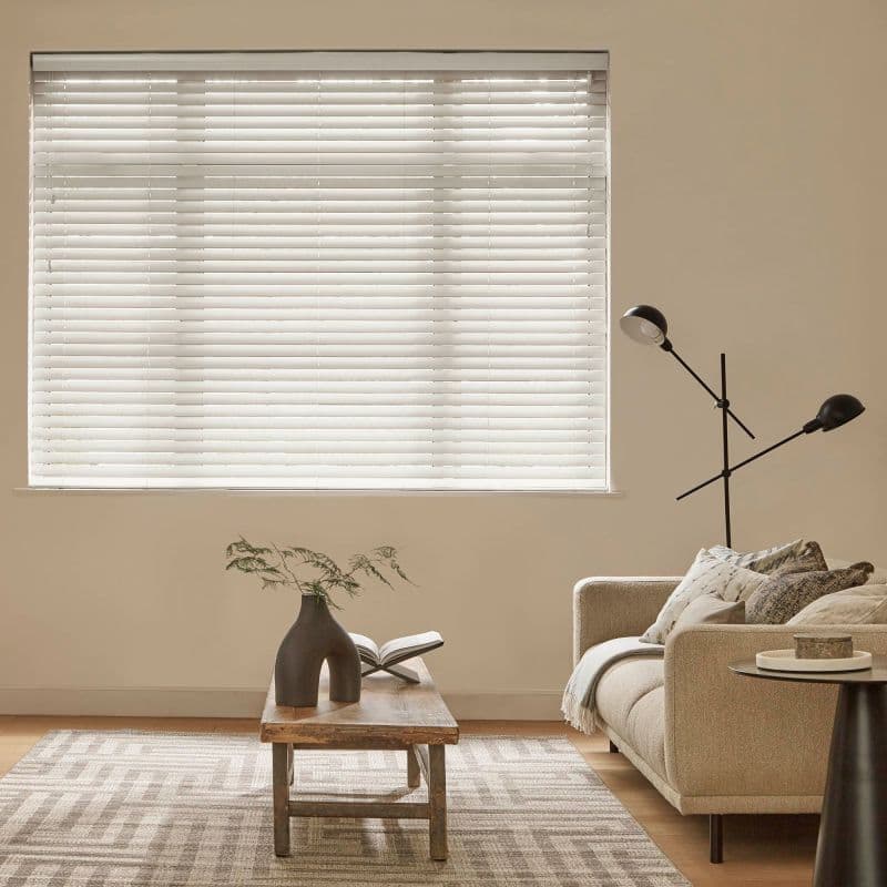 White horizontal Venetian blinds covering a large window, closed and softly diffusing daylight; living room with neutral sofa, wooden coffee table, vase, floor lamp, textured rug and warm ambient light.