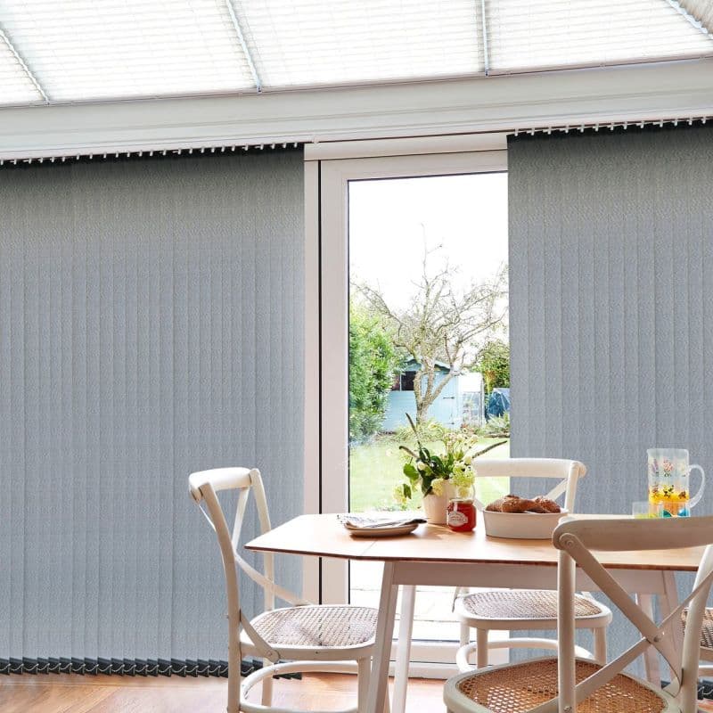 Light grey vertical blinds, floor-to-ceiling and ribbed, mostly closed with a central gap, filtering daylight and revealing a garden through a glass door; small dining table with chairs, vase, pastries.