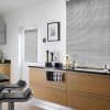 Silver horizontal Venetian blinds covering a large window and adjacent glazed door, thin metallic slats fully lowered; slats angled, filtering diffuse daylight; modern kitchen with light‑wood cabinets, black countertop, stools.