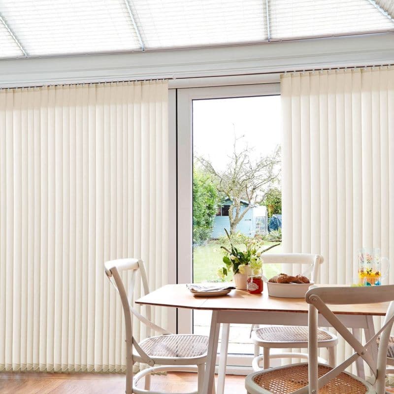 Beige vertical blinds covering full-height sliding glass doors, partially parted to reveal central pane; filtering soft daylight into a bright dining nook with wooden table, chairs, and garden view.