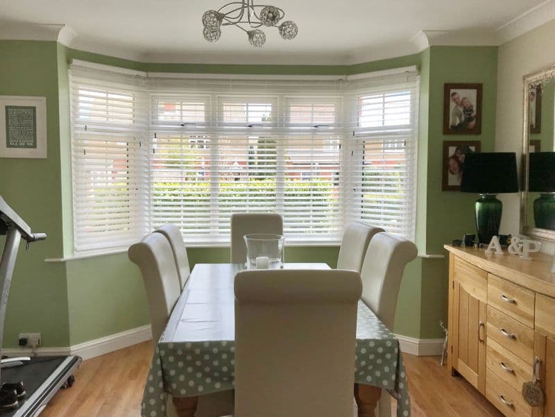 White horizontal Venetian blinds covering a bay window, partly closed and filtering daylight; a dining table with cream chairs faces them in a pale-green room with a wooden sideboard.
A & P