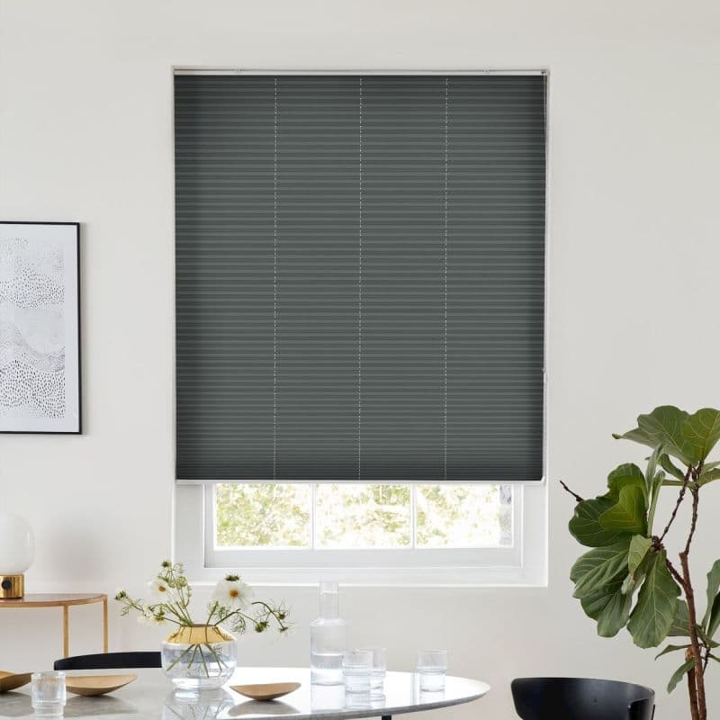 Dark gray pleated cellular shade lowered over a window, subtle vertical seams blocking most light while leaving a slim bright gap above a dining table with vase and potted plant.