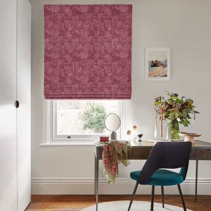 Textured rose‑pink Roman blind (lowered, softly folded) covering window and filtering daylight; small home office with wooden desk, teal chair, vase of flowers, neutral walls and garden view.