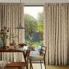 Printed, pleated, floor-length curtains with a beige-and-white leaf pattern, drawn slightly apart to frame a sliding glass door; allowing daylight into a cozy dining area with a set table and garden view.