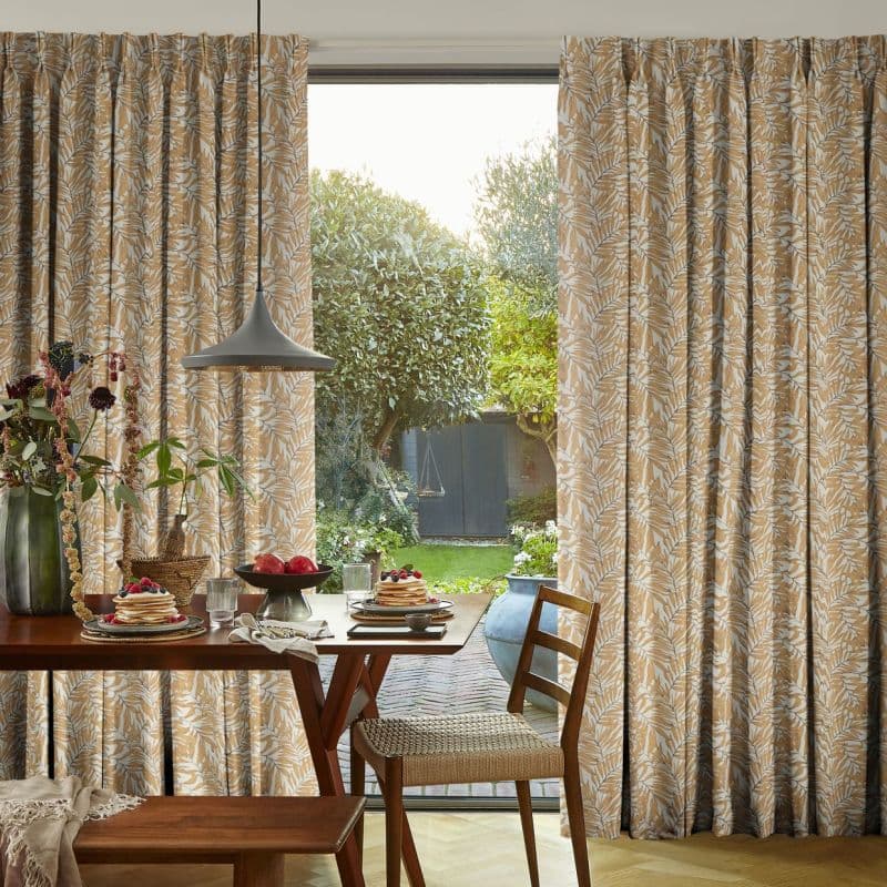 Printed, pleated, floor-length curtains with a beige-and-white leaf pattern, drawn slightly apart to frame a sliding glass door; allowing daylight into a cozy dining area with a set table and garden view.