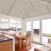 White pleated cellular blinds: roof panels fully lowered, window and door blinds partly lowered, filtering light into a bright conservatory with wooden table, chairs and patio-garden view.