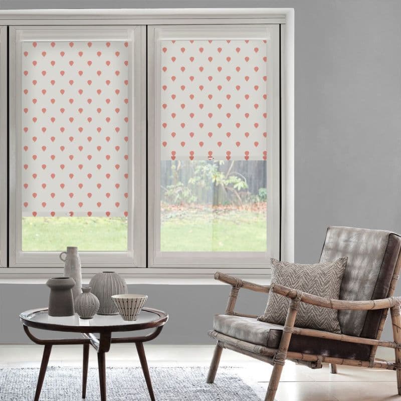 Patterned roller blinds with coral teardrop motifs — left lowered, right half‑raised — filtering daylight on a double window; living room with rattan armchair, side table, grey walls, garden beyond.