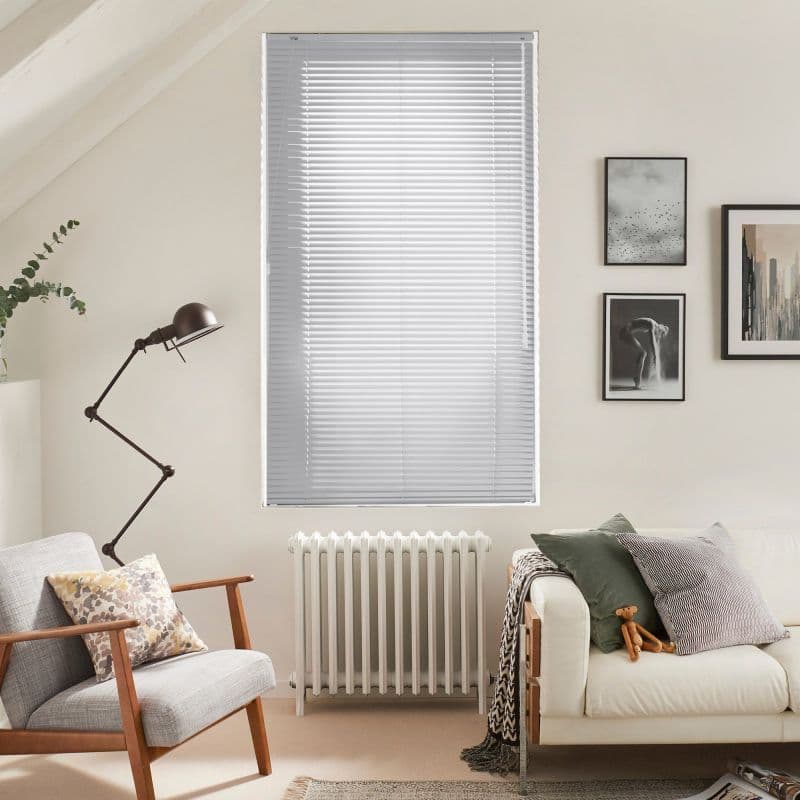 White horizontal venetian blinds fully lowered and closed, softly filtering daylight; set in a neutral-toned living room above a radiator, flanked by an armchair, lamp, sofa and framed art.