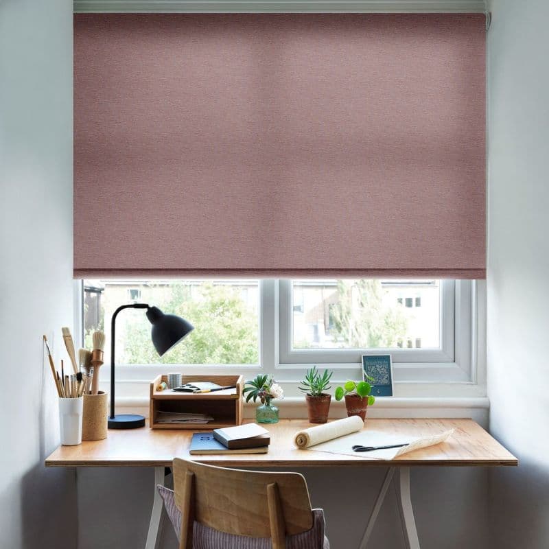 A fabric roller blind, dusty-rose, fully lowered with a narrow bottom gap, blocking and softly diffusing daylight; tidy wooden desk beneath with lamp, potted plants, brushes and notebooks.