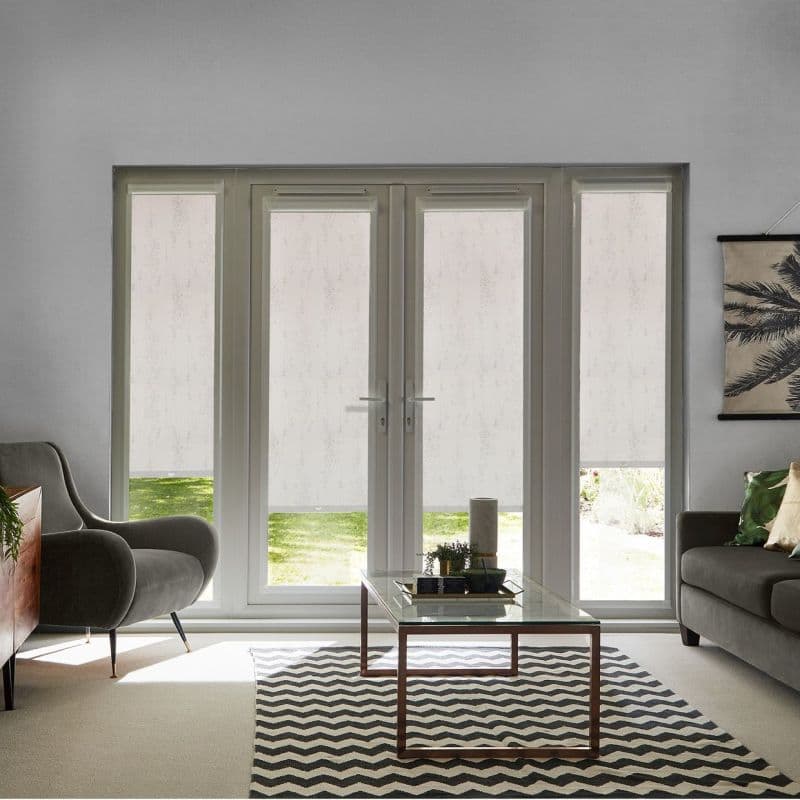 White patterned roller blinds on central French doors and sidelights, lowered to filter daylight and reveal a lawn strip beneath; living room with grey seating, coffee table and chevron rug.
