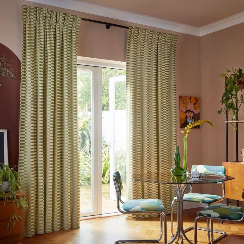 Floor-length green-and-cream checkered curtains on a rod over patio doors, drawn aside, filtering light and framing an open glass door in a sunny dining nook with table, chairs and plants.