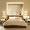 Beige horizontal Venetian blinds covering recessed window, lowered and tilted to filter soft daylight; beige double bed centered beneath, flanked by mirrored bedside tables and lit lamps in neutral-toned bedroom.