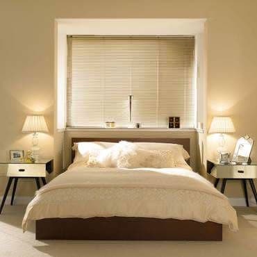 Beige horizontal Venetian blinds covering recessed window, lowered and tilted to filter soft daylight; beige double bed centered beneath, flanked by mirrored bedside tables and lit lamps in neutral-toned bedroom.