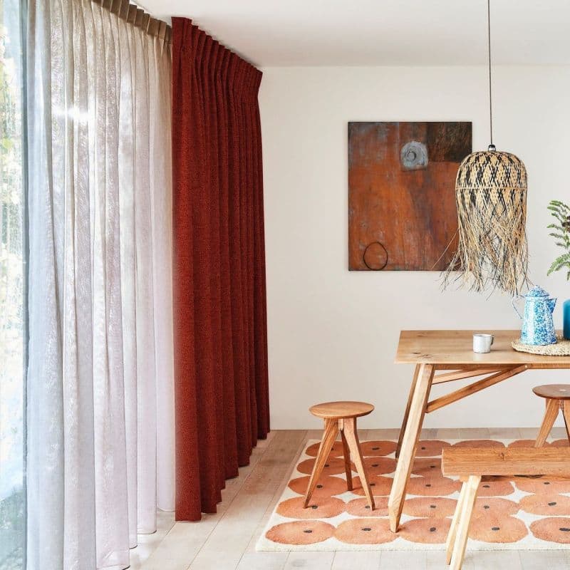 Floor-to-ceiling sheer white panels with pleated rust-red drapes at left, rust curtain drawn while sheer filters daylight; dining nook with wooden table, stools, woven pendant, abstract wall art, patterned rug.