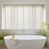 Cream horizontal slatted blinds with vertical ladder tapes cover a recessed window, closed and softly filtering daylight, above a freestanding white bathtub in a neutral, plant‑accented bathroom.